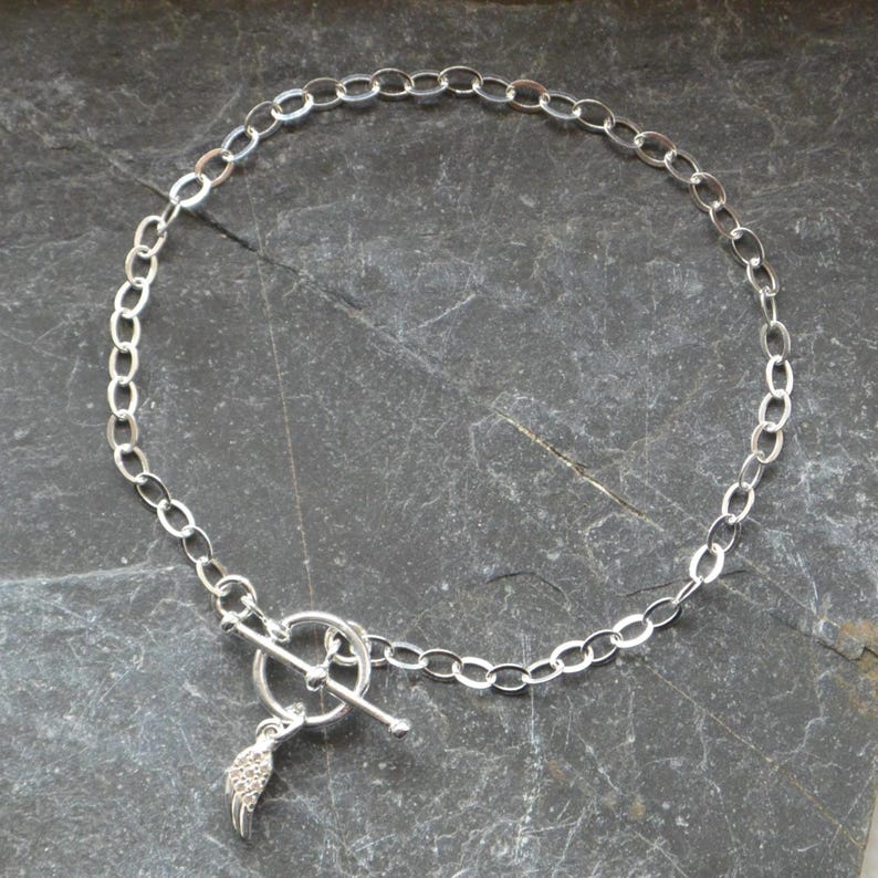 Pu&ograve; includere: Una collana color argento con una catena a maglie ovali e una chiusura a leva. Un piccolo ciondolo a forma di ala &egrave; appeso alla chiusura. La collana &egrave; presentata su una superficie di pietra grigio scuro.