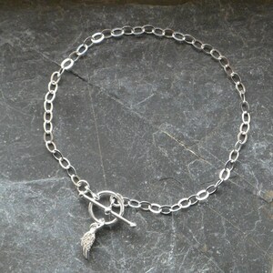 Pu&ograve; includere: Una collana argentata con una catena a maglie ovali e una chiusura a toggle. Un piccolo ciondolo a forma di ala pende dalla chiusura. Il gioiello &egrave; esposto su una superficie scura e strutturata.