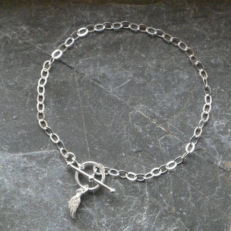 Pu&ograve; includere: Una collana argentata con una catena a maglie ovali e una chiusura a toggle. Un piccolo ciondolo a forma di ala pende dalla chiusura. Il gioiello &egrave; esposto su una superficie scura e strutturata.