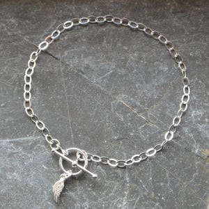 Pu&ograve; includere: Una collana color argento con una catena a maglie ovali e una chiusura a toggle. Un piccolo ciondolo a forma di ala pende dalla chiusura. La collana &egrave; esposta su una superficie di pietra grigio scuro.