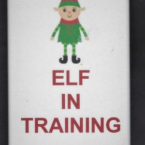 Puede incluir: Un imán blanco con un duende de dibujos animados que lleva un gorro a rayas verde y rojo y pantalones a rayas verde y amarillo. El texto "ELF IN TRAINING" está en letras rojas.