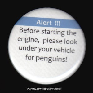 May include: A white button with a blue stripe across the top. The button has the text "Alert!!! Before starting the engine, please look under your vehicle for penguins!" and the website address "www.etsy.com/shop/SwankSpecials".