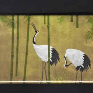 May include: Two white cranes with black wing tips stand in a bamboo forest. The cranes are on a gold background with green bamboo stalks.