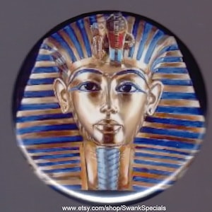 May include: A gold and blue button featuring a stylized image of the pharaoh Tutankhamun's mask. The mask is depicted with a blue and gold headdress and a gold face.