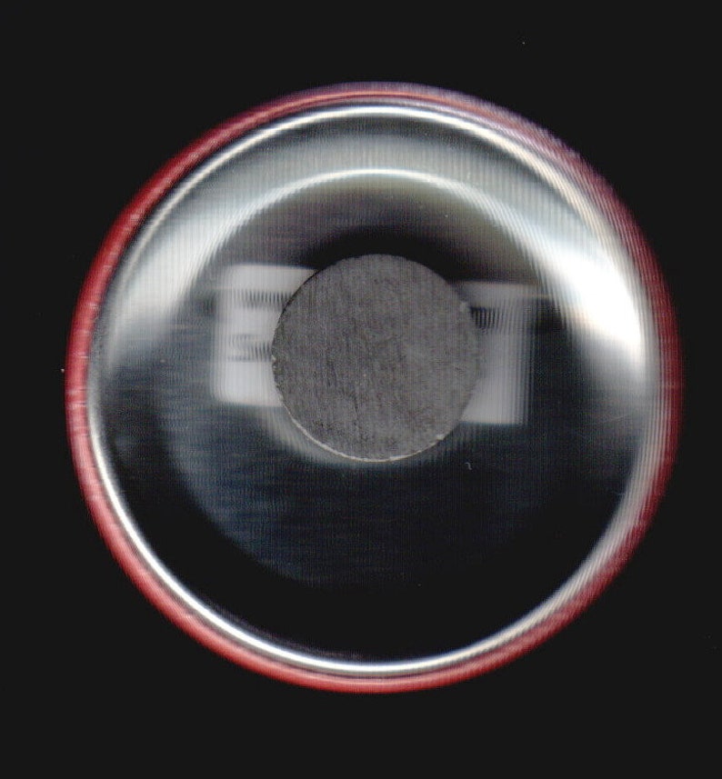May include: Close-up of the back of a round button with a red border. The button has a silver backing with a black circle in the center.