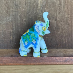May include: A white ceramic elephant figurine with blue and green floral designs. The elephant has its trunk raised and is decorated with yellow flowers.