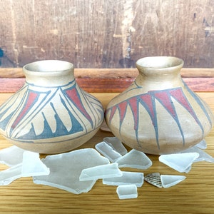 May include: Two small, brown ceramic pots with geometric designs in red, black, and white. The pots are sitting on a wooden surface with pieces of clear glass scattered around them.
