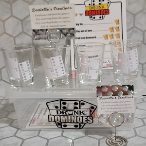 May include: A set of clear shot glasses with black dominoes printed on them. The glasses are arranged in a clear plastic container. A card with the text "DRINK DOMINOES" is visible in the foreground. The card also includes contact information for the creator, Danielle's Creations.