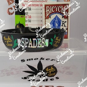 May include: A black ashtray with the word "SPADES" in holographic lettering, a deck of Bicycle playing cards, and a green and black lighter. The image also includes stickers with text and a cannabis leaf.