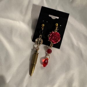 May include: A pair of mismatched earrings. One earring has a gold-coloured bullet charm, clear and black beads. The other earring features a red rose and a red crystal. The earrings are attached to a black display card.