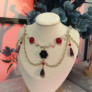 May include: A gold chain necklace with red and white beads, a black flower charm, and dangling red and purple teardrop crystals.
