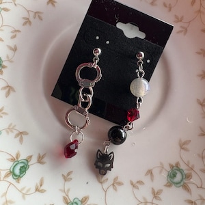 Könnte beinhalten: Ein Paar nicht übereinstimmender Ohrringe. Ein Ohrring hat ein Handfessel-Design mit einem roten Kristalltropfen. Der andere Ohrring hat eine weiße und graue Perle, eine schwarze Perle, einen Wolfskopf-Anhänger und einen roten Kristall. Die Ohrringe sind silberfarben.