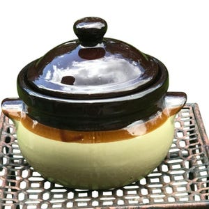 May include: A brown and cream-colored ceramic casserole dish with a lid. The dish has a glossy finish and two handles. The lid has a rounded knob. The dish is sitting on a metal surface.