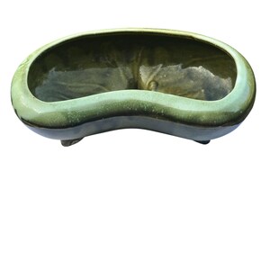 May include: A green ceramic planter with a kidney bean shape. The planter has a glossy finish with a dark green interior and a lighter green exterior. It is supported by three small feet.