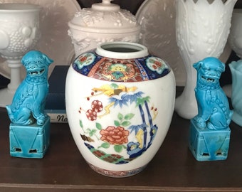Peut inclure: Un vase en porcelaine blanche avec un motif floral et de bambou. Le vase est décoré d'accents rouges, bleus, dorés et verts. Deux chiens foo en céramique bleue sont de chaque côté du vase.