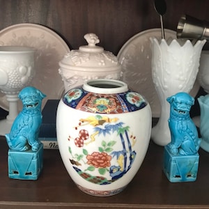May include: A white porcelain vase with a floral and bamboo design. The vase is decorated with red, blue, gold, and green accents. Two blue ceramic foo dogs are on either side of the vase.