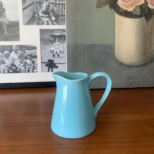 Può includere: Un piccolo brocca in ceramica azzurra con manico. La brocca è su una superficie di legno, con una fotografia in bianco e nero incorniciata e un dipinto di rose rosa sullo sfondo.