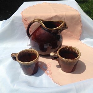 May include: A brown glazed ceramic pitcher and two matching mugs on a white tablecloth with a pink doily.