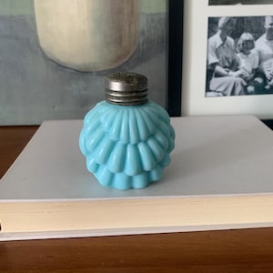 May include: A light blue, antique glass salt shaker with a ruffled, layered design. The top is a silver-colored metal with small holes. The shaker is sitting on a white book.