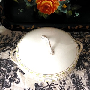 Puede incluir: Una fuente de cerámica blanca con tapa, con ribete dorado y un delicado diseño de hojas verdes. El plato tiene dos asas y un asa curva en la tapa. El fondo incluye una bandeja negra con obras de arte florales y una tela estampada.