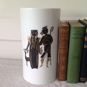May include: White ceramic vase with a black and gold design of a couple in silhouette. The vase is sitting on a table next to a stack of books.