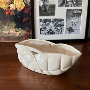 May include: Cream-colored ceramic leaf-shaped planter. The planter has a glossy finish and is resting on a wooden surface. The planter has a large opening and is designed with leaf-like details. A framed picture is in the background.