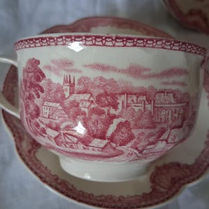 May include: A white teacup and saucer set with a red transferware design depicting a village scene. The cup has a curved handle and sits on a matching saucer with a scalloped edge. The rim of the cup has a decorative border.