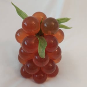 May include: A cluster of artificial grapes with translucent, reddish-brown orbs. The grapes are arranged in a bunch, with green leaves attached to the stem. The grapes have a glossy finish.