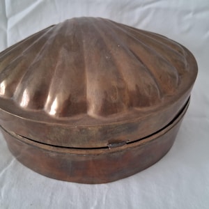 May include: A vintage, copper-toned metal box shaped like a scallop shell. The lid and base are oval, with the lid featuring a ribbed, fan-like design. The box is closed with a small metal clasp. The box is on a white surface.