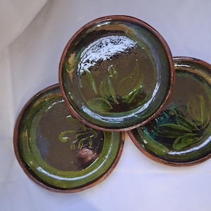 May include: Three handmade ceramic plates with a dark green glaze and brown rims. Each plate features a painted leaf design in shades of green. The plates are arranged on a white surface.