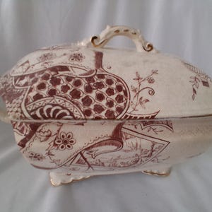 May include: Cream-colored, antique serving dish with a lid. The dish features a brown floral and landscape pattern. It has a handle on the lid and two side handles. The dish is on small feet with gold trim.