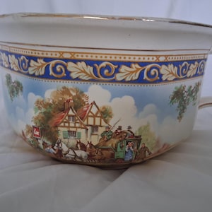 May include: A ceramic chamber pot featuring a colorful transferware scene of a horse-drawn carriage arriving at a half-timbered inn. The rim is accented with gold trim and a blue band with gold leaf patterns. It includes a single side handle.