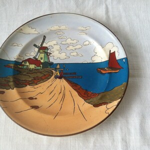 May include: A decorative plate featuring a coastal scene with a windmill, red-sailed boat, and a pier. The plate has a brown rim and a light blue sky with white clouds. The artwork depicts a path leading to the pier.