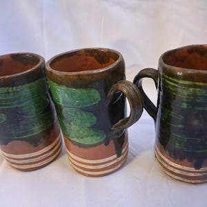 May include: Three handmade ceramic mugs. Each mug features a cylindrical shape with a dark brown rim and handle. The mugs have a green and brown painted design with a cream-colored base and horizontal stripes.