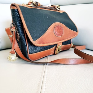 May include: A black and brown leather shoulder bag with a flap closure and a brass buckle. The bag has a long adjustable strap and a small circular charm with a logo.