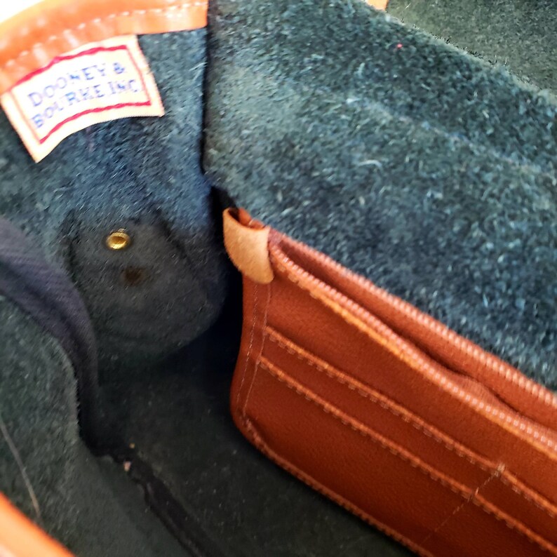 May include: Inside view of a brown leather handbag with a zippered pocket. The interior is lined with dark green fabric and has a Dooney & Bourke Inc. label.