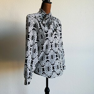 May include: A white blouse with a black and white geometric pattern. The blouse has a high neck and a tie at the neck.