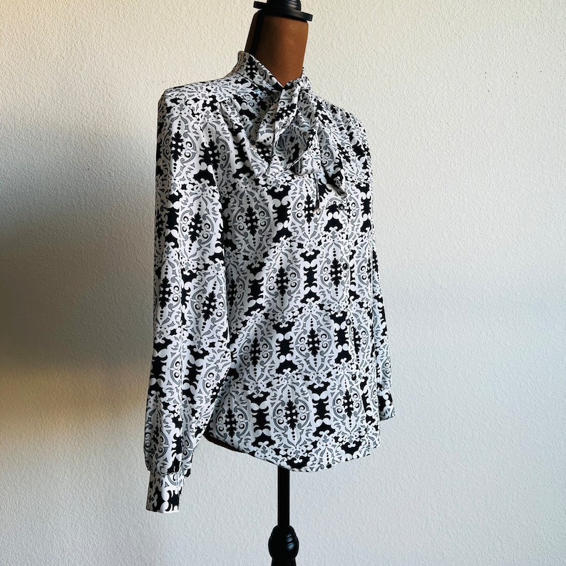 May include: A white blouse with a black and white geometric pattern. The blouse has a high neck and a tie at the neck.