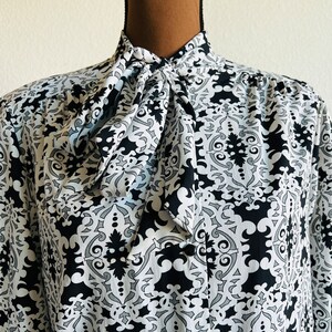 May include: A black and white patterned blouse with a large bow tie at the neck. The pattern is a repeating geometric design.