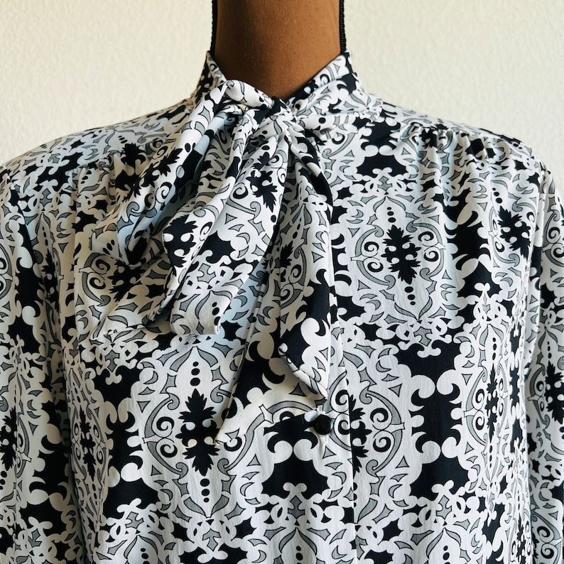 May include: A black and white patterned blouse with a large bow tie at the neck. The pattern is a repeating geometric design.