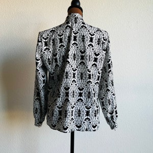 May include: A white long-sleeved blouse with a black and white geometric pattern. The blouse has a high neck and long sleeves with a gathered cuff.