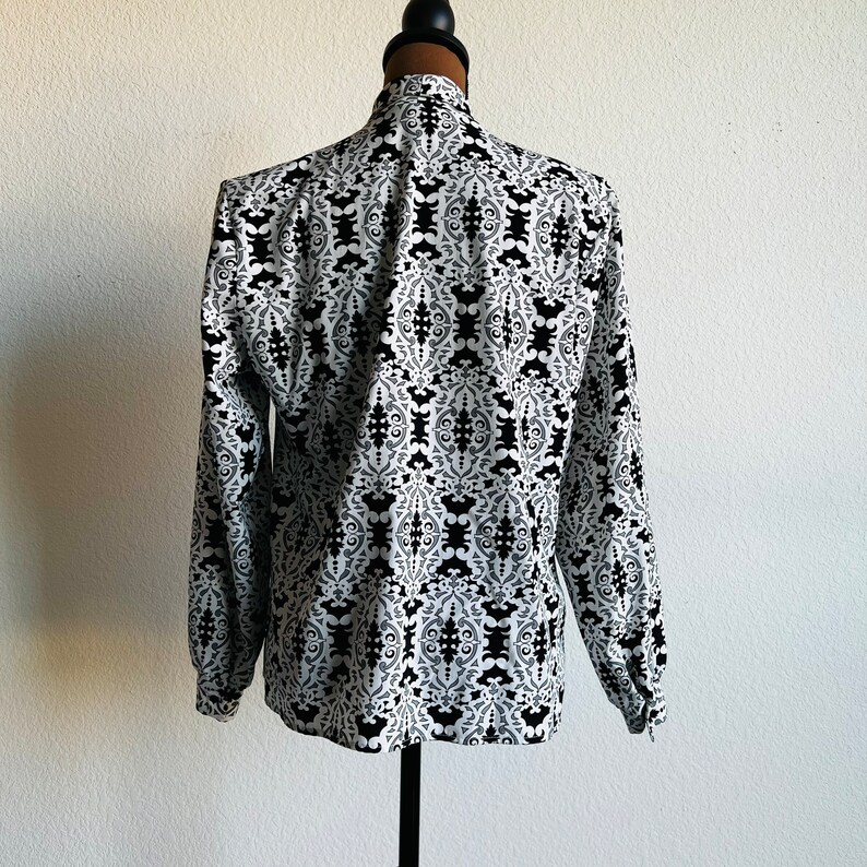 May include: A white long-sleeved blouse with a black and white geometric pattern. The blouse has a high neck and long sleeves with a gathered cuff.