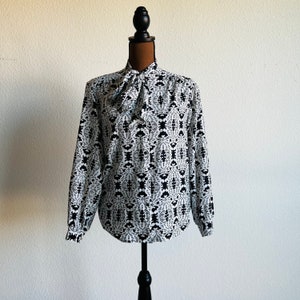 May include: A white blouse with a black and white geometric pattern. The blouse has a high neck and a bow tie at the neck.