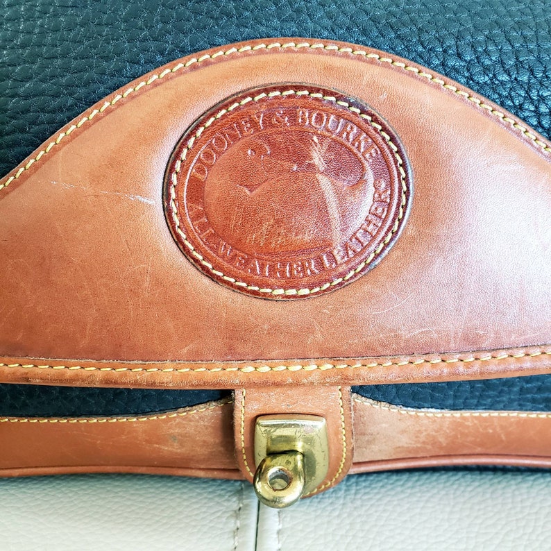 May include: A brown leather Dooney & Bourke bag with a black leather trim and a brass clasp. The bag has a stitched design and a circular logo that reads "Dooney & Bourke All Weather Leather".