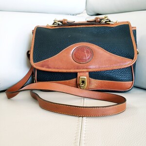 May include: A black and brown leather satchel with a long brown strap. The bag has a brass buckle and a circular logo with the text "Dooney & Bourke" and "All Weather Leather".