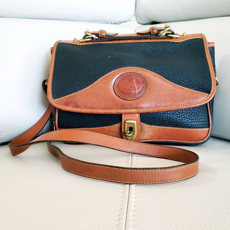 May include: A black and brown leather satchel with a long brown strap. The bag has a brass buckle and a circular logo with the text "Dooney & Bourke" and "All Weather Leather".