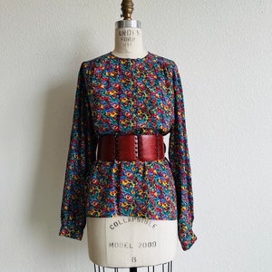 May include: A long-sleeved blouse with a colorful floral print and a brown leather belt with metal accents. The blouse has a round neckline and a relaxed fit.