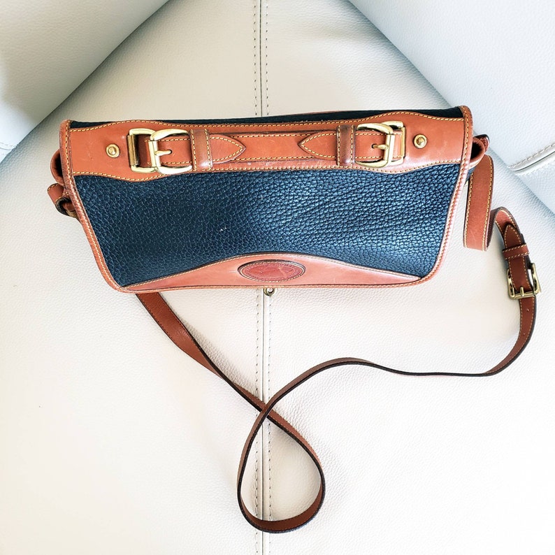 May include: A black leather shoulder bag with brown leather trim and gold hardware. The bag has a flap closure with two buckles and a long adjustable strap.