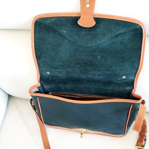 May include: A black leather satchel with brown leather trim and a green suede lining. The bag has a flap closure with a brass buckle and a long adjustable shoulder strap.