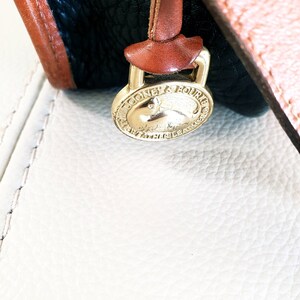 May include: Close-up of a gold-colored Dooney & Bourke brand tag with a small, embossed rabbit logo. The tag is attached to a brown leather strap.